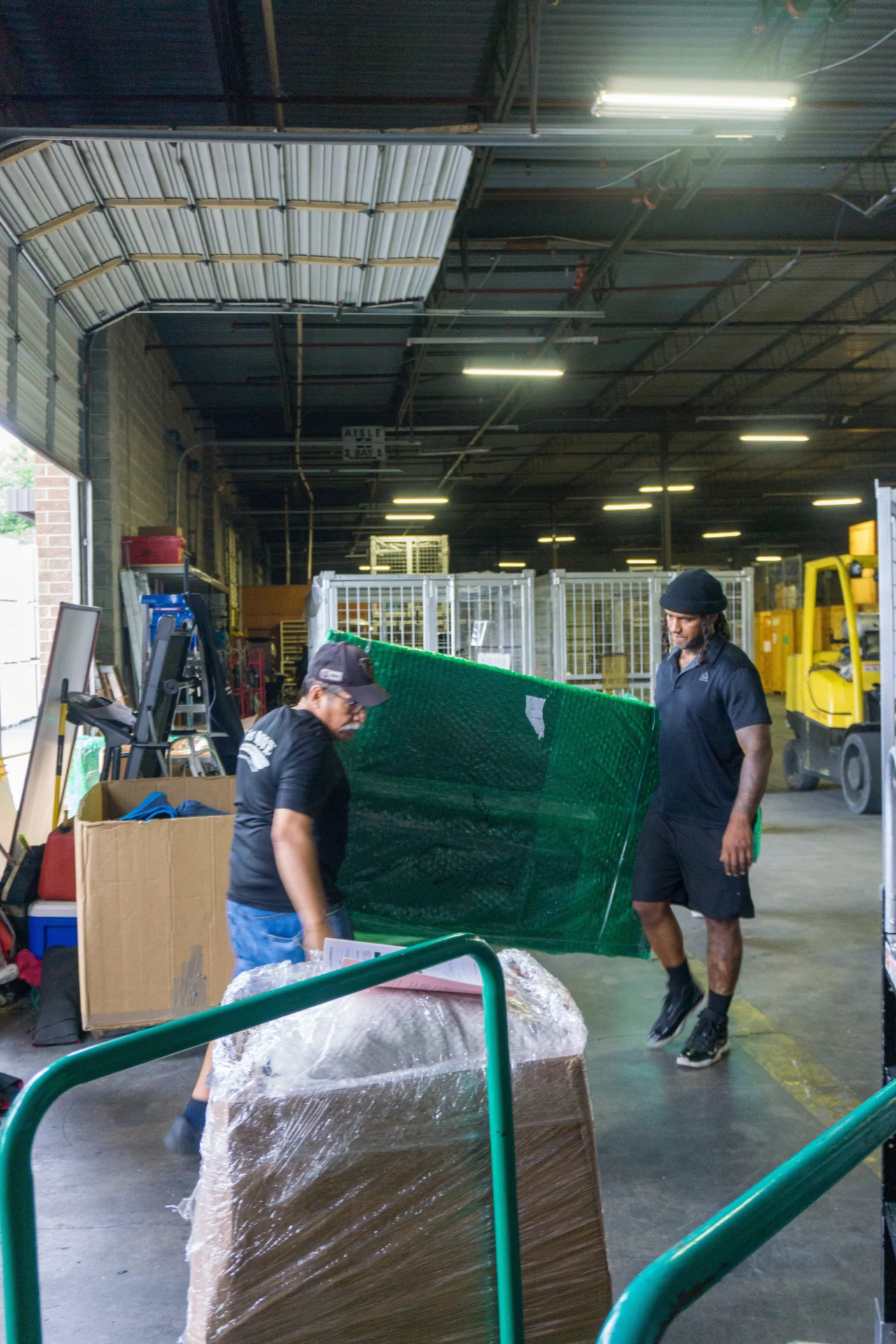 Warehouse workers moving green tarp-covered item.